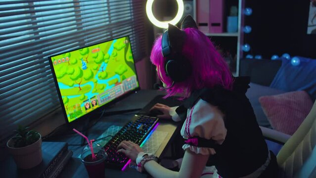 Teen girl wearing maid cosplay dress with pink wig, cat ears and gaming headset playing online game on computer