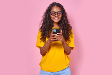 Young happy Indian woman teenager with smile using phone and chatting with friends from school exchanging funny pictures or reading comments in social network stands on isolated pink background.