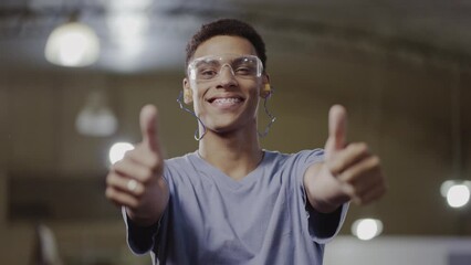 young studant Brazilian man with short hair wearing safety glasses smiling at the camera and giving the thumbs up sign in a factory in Brazil