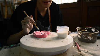 Cropped shot of man painting clay plate with a paintbrush at desk in ceramic workshop