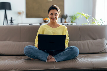 Beautiful young woman working with her laptop while sitting on a couch at home.