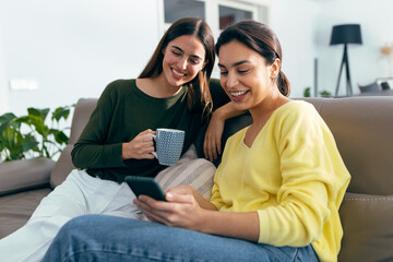 Two smiling young women talking while watching smartphone sitting on couch in the living room at home.