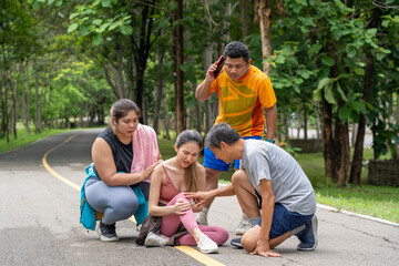 A young woman in fitness clothes with knee pain grabbing her knee with both hands while sitting on the middle of a running track at a local park with three other runners gather around to help