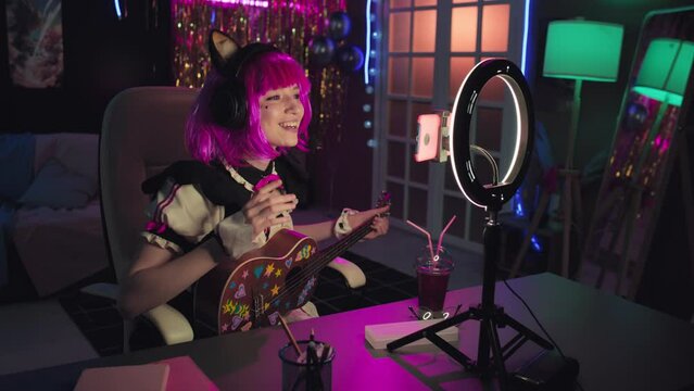 Caucasian girl wearing neko maid costume with pink wig holding ukulele decorated with stickers making content for vlog