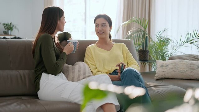 Video of two smiling young women talking while drinking coffee sitting on couch in the living room at home.