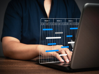 Fototapeta premium Project manager working on a laptop and updating tasks and milestones progress planning with a Gantt chart scheduling interface on a virtual screen.