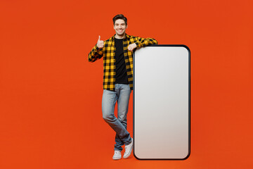 Full body smiling happy young man in yellow checkered shirt black t-shirt big huge blank screen mobile cell phone smartphone with workspace area show thumb up isolated on plain red orange background