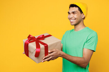 Side view young man of African American ethnicity wears casual clothes green t-shirt hat hold in hand present box with gift ribbon bow look aside isolated on plain yellow background Lifestyle concept.