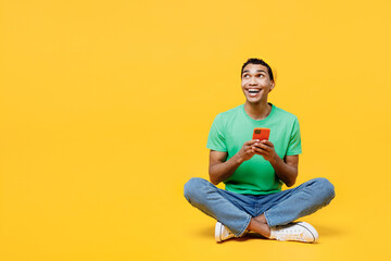 Full body young man of African American ethnicity he wears casual clothes green t-shirt hat sitting hold in hand use mobile cell phone look aside isolated on plain yellow background Lifestyle concept