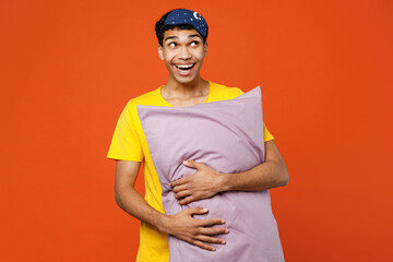 Calm young smiling happy fun man wear pyjamas jam sleep eye mask rest relax at home hold pillow look overhead on area isolated on plain orange background studio portrait. Good mood night nap concept.