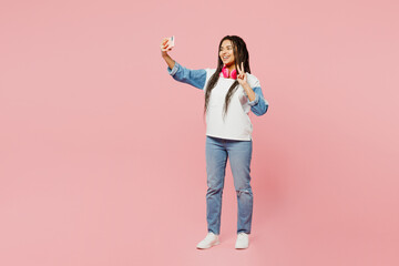 Full body young woman of African American ethnicity wear white sweatshirt casual clothes doing selfie shot on mobile cell phone show v-sign isolated on plain pastel pink background. Lifestyle concept.