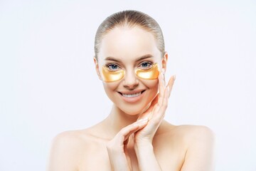 Attractive woman touches face, smiles, applies golden hydrogel eye patches, isolated on white...