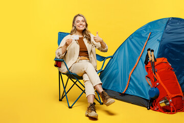 Full body young woman sit near bag with stuff tent hold credit bank card show thumb up like gesture isolated on plain yellow background Tourist walk on spare time Hiking trek rest travel trip concept