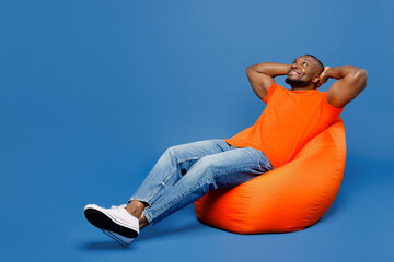 Full body young man of African American ethnicity he wear orange t-shirt sit in bag chair hold...