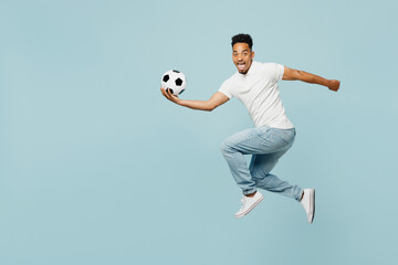 Full body side profile view of young man fan wearing t-shirt cheer up support football sport team jump high hold catch soccer ball watch tv live stream isolated on plain pastel blue color background.