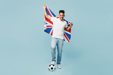 Full body young overjoyoed happy man fan wearing basic t-shirt cheer up support football sport team hold British flag soccer ball watch tv live stream isolated on plain pastel blue color background.