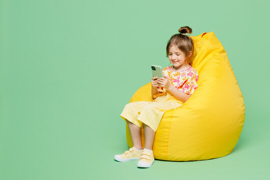 Full Body Little Child Kid Girl 6-7 Years Old Wearing Casual Clothes Sit In Bag Chair Hold In Hand Use Mobile Cell Phone Isolated On Plain Green Background Mother's Day Love Family Lifestyle Concept.