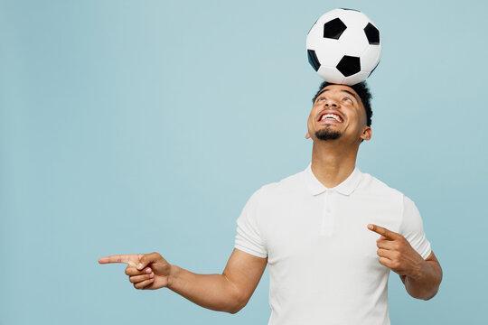 Young fun cool man fan wearing basic t-shirt cheer up support football sport team holding in hand soccer ball on head point aside watch tv live stream isolated on plain pastel blue color background.