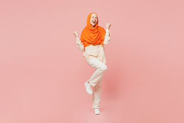 Fototapeta na wymiar Full body overjoyed young arabian asian muslim woman wear orange abaya hijab do winner gesture clench fist raise up leg isolated on plain pink background. Uae middle eastern islam religious concept.