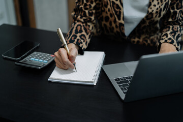 Businesswoman sitting at a table at home working on a laptop and writing down ideas in a notebook.