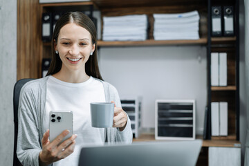 Woman sits in a office working on smartphone play social media and enjoys a coffee.