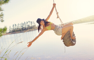 Lake, rope swing and a woman in nature sitting on a tyre for carefree fun or adventure during...