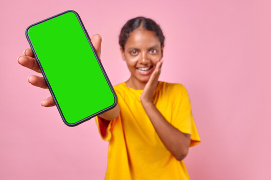 Young positive casual Indian woman teen stretches hand with smartphone to camera showing off received message with invitation to become university student stands in pink studio. Green screen - Powered by Adobe