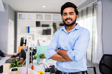 Successful Indian businessman entrepreneur in casual smiling and looking at the camera while standing with his arms crossed. Happy businessman standing in the office of a modern corporate workplace.