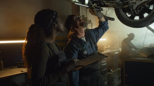 Mature Caucasian auto technician holding torch and his Black female co-worker with paper on clipboard inspecting car underbody