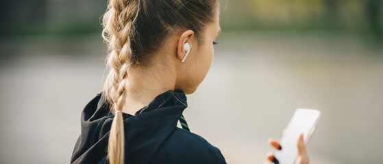 Young smiling girl making sport and running in the park using her phone to listen the music with wireless headphones on sunset in the city watching the screen