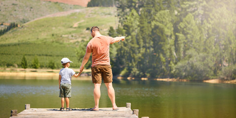 Fototapeta na wymiar Back, lake and a father holding hands with his son while looking at the view while outdoor in nature together. Love, family or kids with a man and boy child standing on a pier for bonding by water