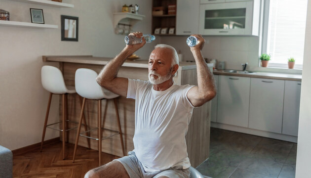 Bearded senior man doing exercises with water bottles sitting on fitball stretching at home indoor.