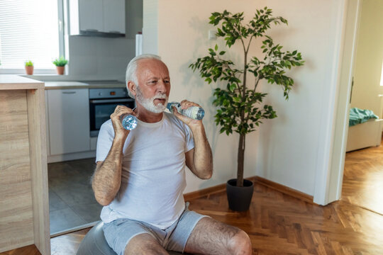 Bearded senior man doing exercises with water bottles sitting on fitball stretching at home indoor.