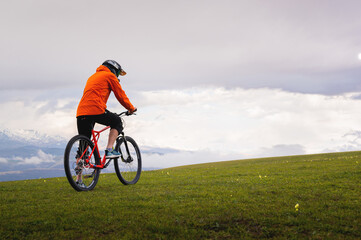 man stands with a bicycle on a green lush meadow, against the backdrop of mountains and a cloudy...