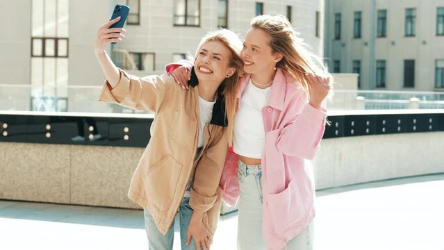 Two young beautiful blond smiling hipster women in trendy summer clothes. Carefree models posing on street background. Positive models having fun. Holding smartphone, take selfie photos