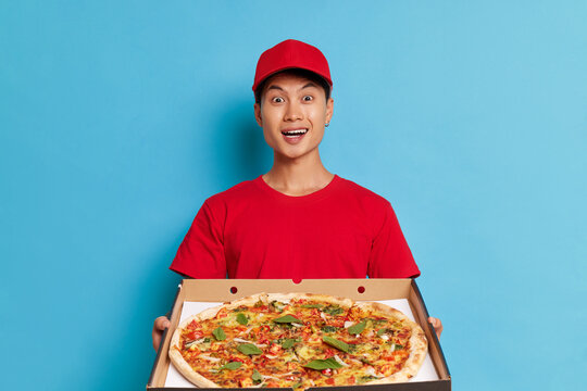 Surprised asian delivery man in red uniform holding pizza and looking at camera isolated over blue background
