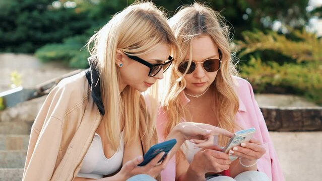 Two young beautiful hipster female. Carefree women sitting at the stairs in the street in sunglasses. Models having fun. They looking at cellphone screen, using mobile apps, hold smartphone 