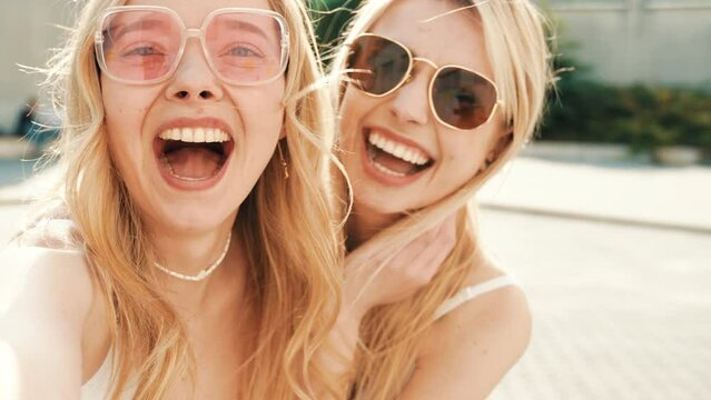 Two young beautiful blond smiling hipster women in trendy summer white t-shirt and jeans clothes. Carefree women posing in street. Positive models having fun in sunglasses. Taking Pov selfie photos