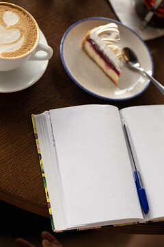 A mug of cappuccino, a slice of cake and a notebook on the table in a cafe.