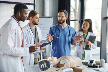 multicultural students in white coats writing near healthcare worker gesturing and talking next to CPR manikin and medical equipment on first aid seminar, emergency situations response concept