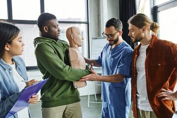 medical instructor assisting african american man practicing life-saving techniques in case of choking on CPR manikin near multiethnic participants, emergency situations preparedness concept