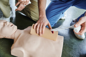 high angle view of medical instructor tamponing wound on simulator with bandage near CPR manikin and young man during first aid training seminar, emergency response concept, cropped view