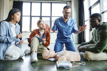 professional paramedic gesturing and talking to multiethnic participants near CPR manikin and...