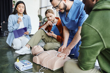 first aid seminar, medical instructor applying defibrillator pads on CPR manikin near multiethnic team with clipboard and notebook in training room, health care and life-saving techniques concept - Powered by Adobe