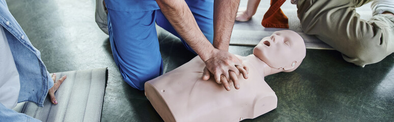 first aid seminar, hands-on learning, cropped view of paramedic showing chest compressions on CPR manikin near young participants in training room, life-saving skills and techniques concept, banner © LIGHTFIELD STUDIOS