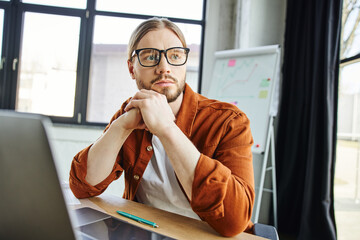 stylish and pensive businessman in eyeglasses and trendy casual clothes looking away near laptop and pen on work desk next to flip chart with graphs on blurred background in modern office
