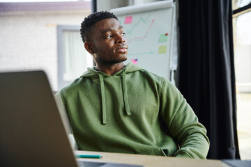 young african american entrepreneur in green hoodie sitting and looking away while planning startup near flip chart with business analytics in modern office, professional development