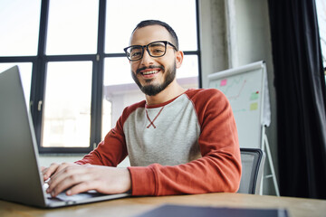 youthful and smiling bearded businessman in eyeglasses typing on laptop and looking at camera in front of flip chart with graphs on blurred background in contemporary office