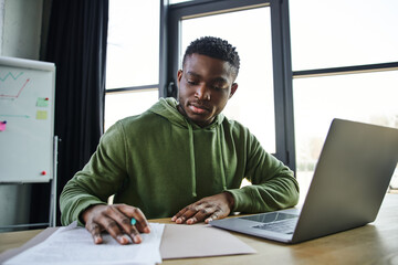 young and stylish african american businessman sitting at workplace and working with documents near laptop and flip chart on blurred background in office, ambitious and career oriented youth