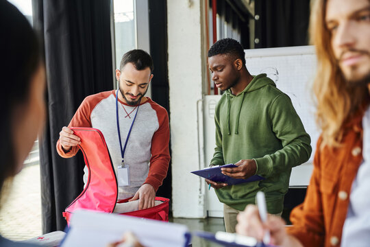 medical training and first aid seminar, bearded paramedic showing first aid kit to african american man with clipboard near young participants on blurred foreground, emergency response concept